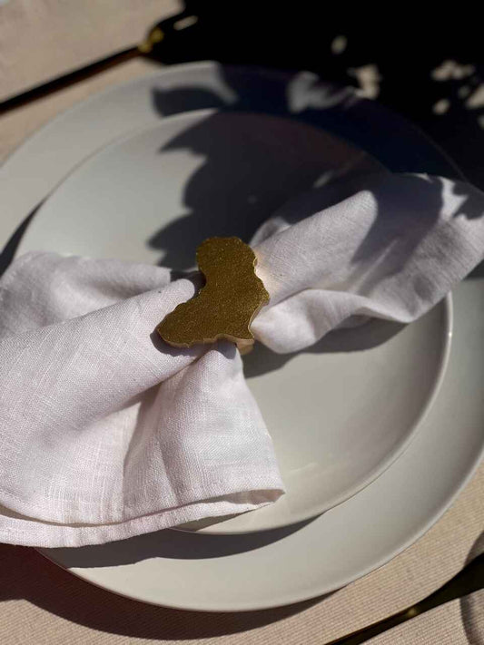 Africa napkin ring in gold on an off white plate, glass of water and gold cutlery, on a cream placemat.