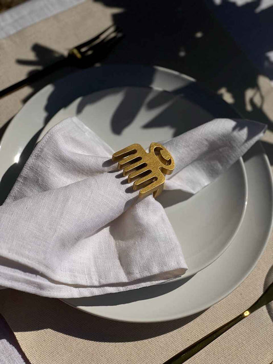Dining table setting with off white plate, folded napkin with a gold Duafe adinkra napkin ring, gold cutlery and a glass of water on a beige tablecloth.