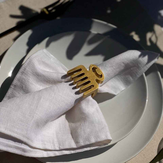 Dining table setting with off white plate, folded napkin with a gold Duafe adinkra napkin ring, gold cutlery and a glass of water on a beige tablecloth.
