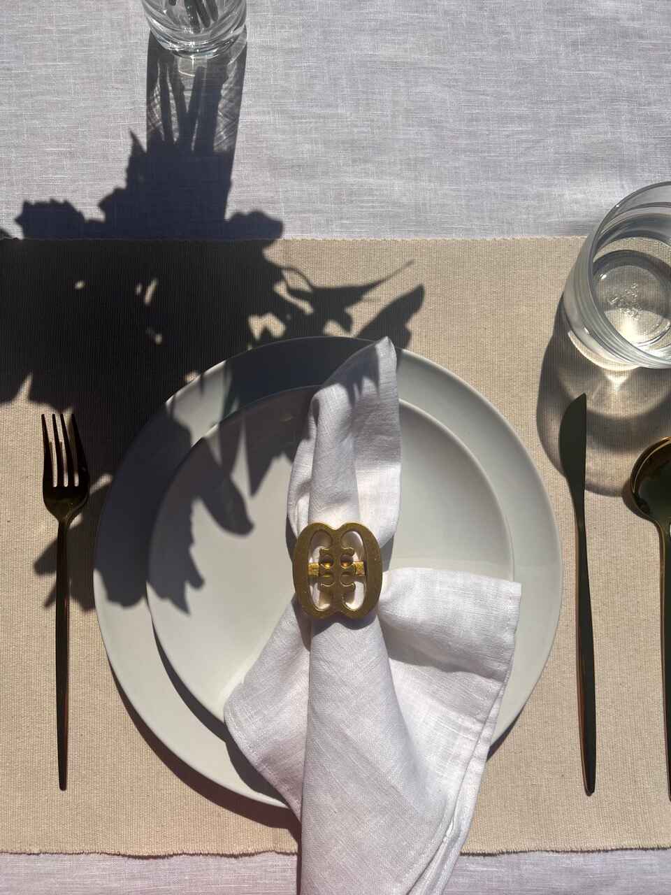 A gold coloured Ese Ne Tekrema adinkra napkin ring on an off white plate, glass of water and gold cutlery.