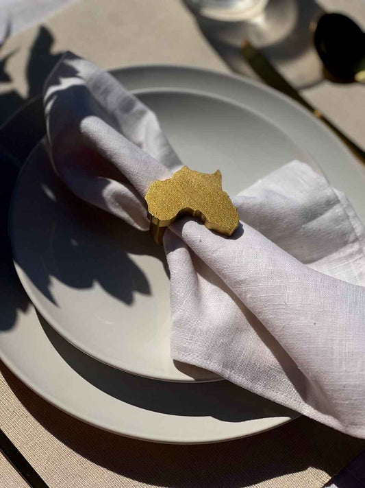 Africa napkin ring in gold on an off white plate, glass of water and gold cutlery, on a cream placemat.