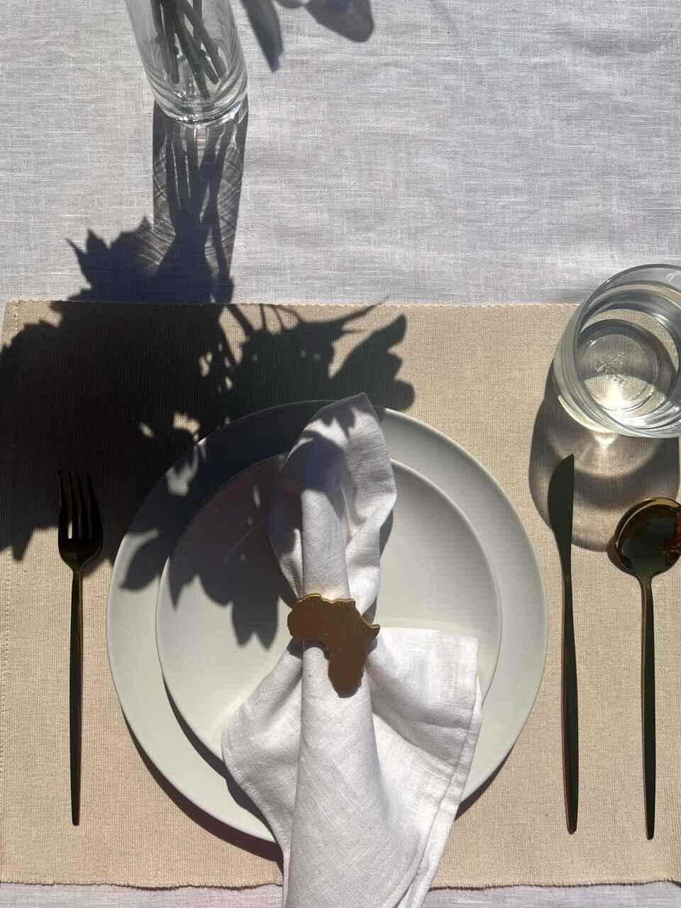 Africa napkin ring in gold on an off white plate, glass of water and gold cutlery, on a cream placemat.