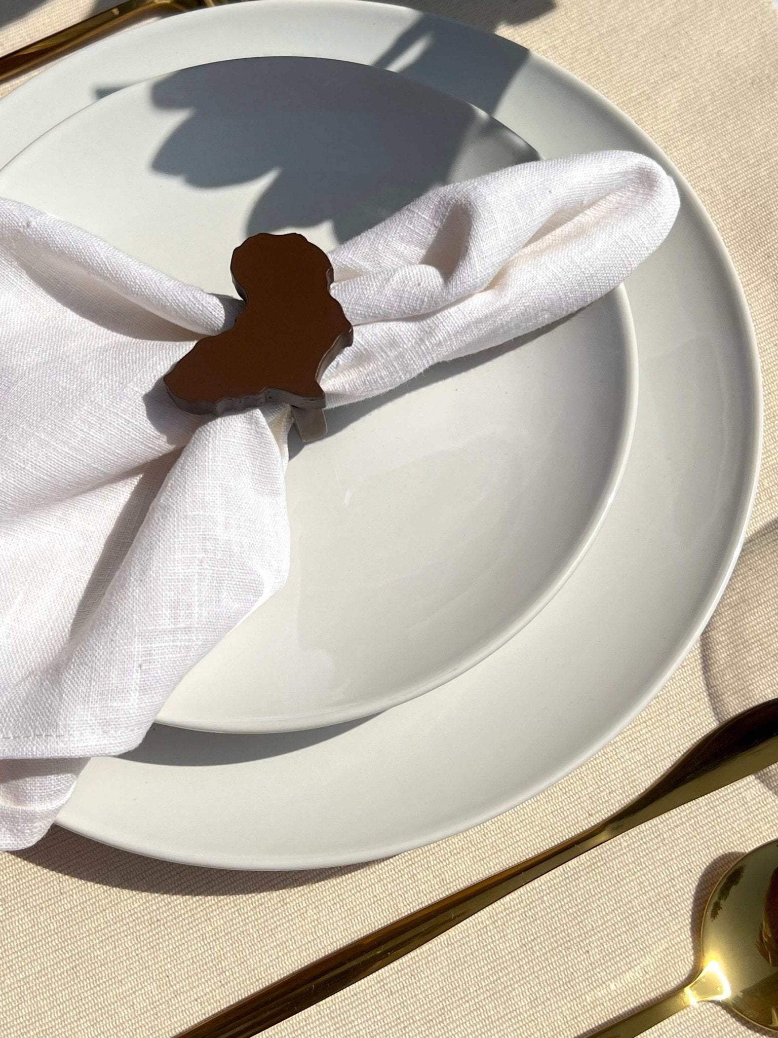 A coffee coloured Africa napkin ring on an off white plate with gold cutlery.
