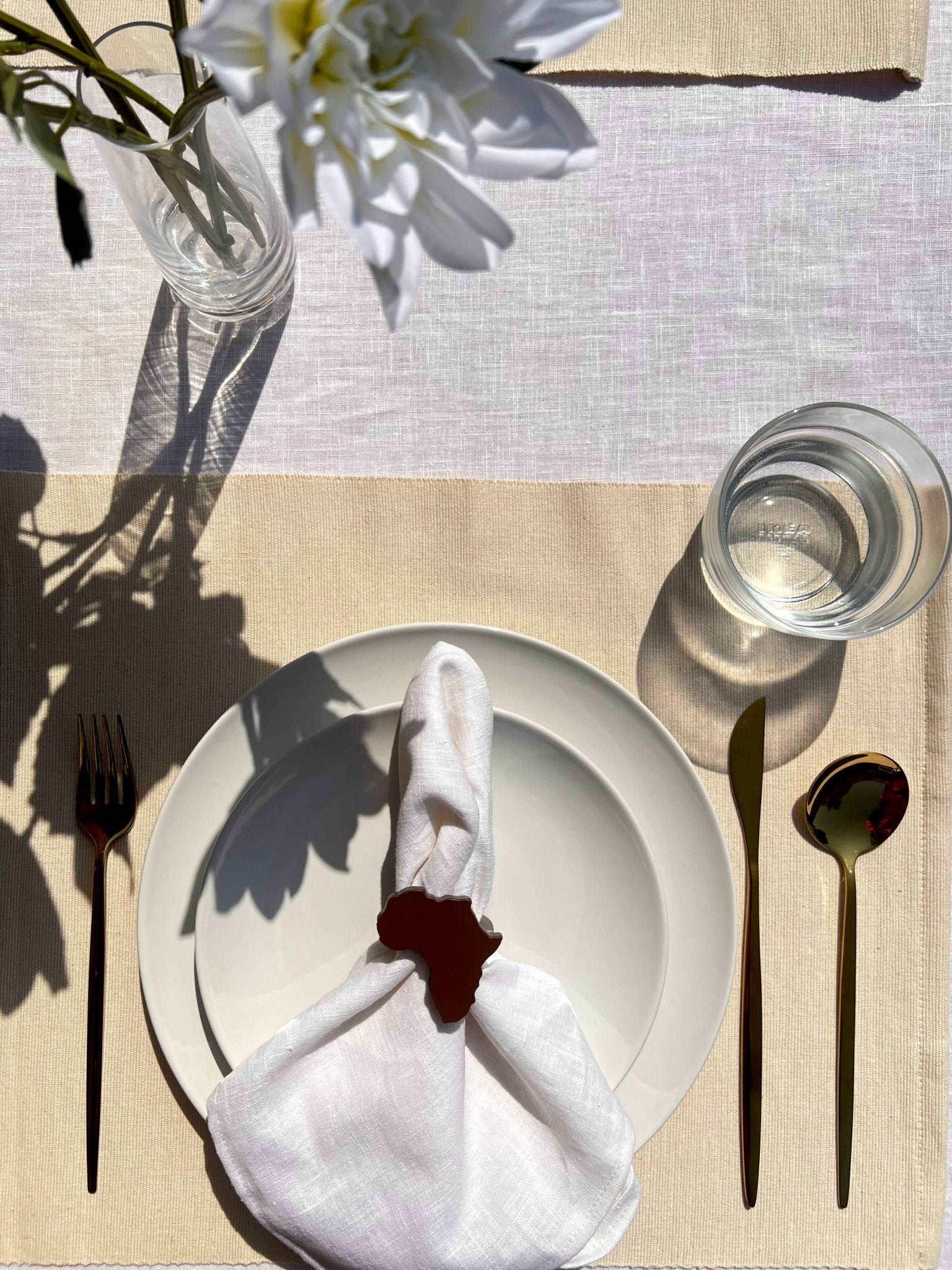 Dining table setting with off white plate, folded napkin with a coffee coloured Africa shaped napkin ring, gold cutlery and a glass of water.