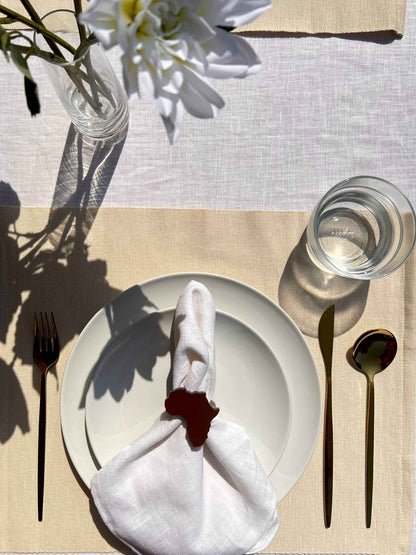 Dining table setting with off white plate, folded napkin with a coffee coloured Africa shaped napkin ring, gold cutlery and a glass of water.