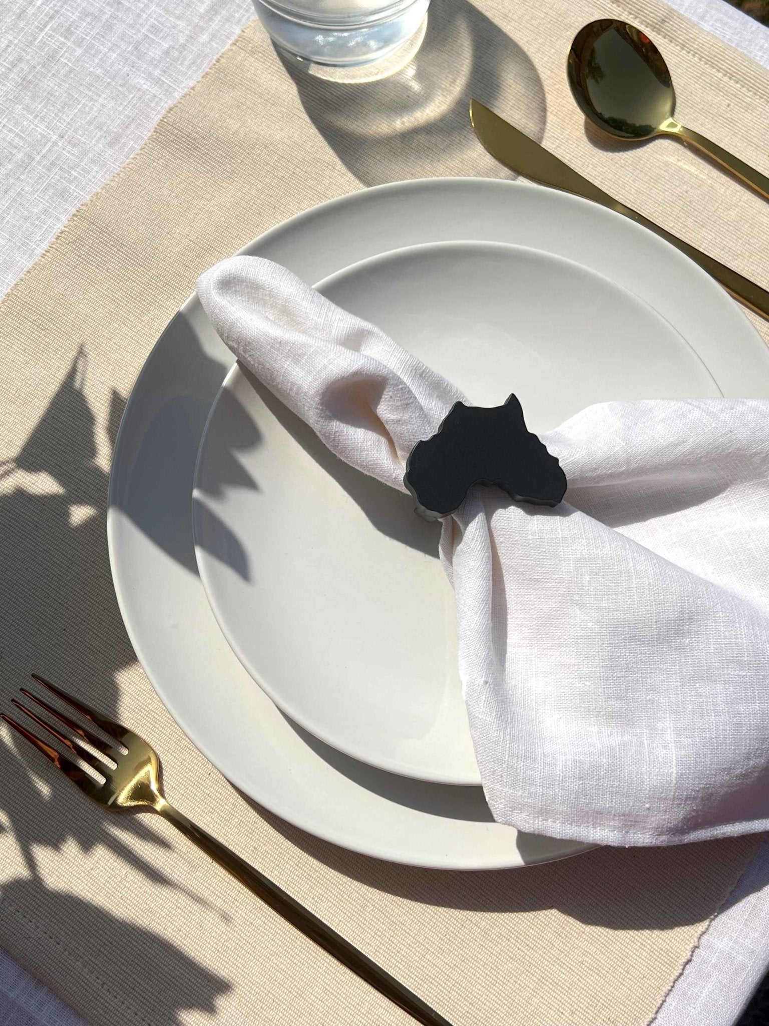 Africa napkin ring in black on an off white plate, glass of water and gold cutlery, on a cream placemat.