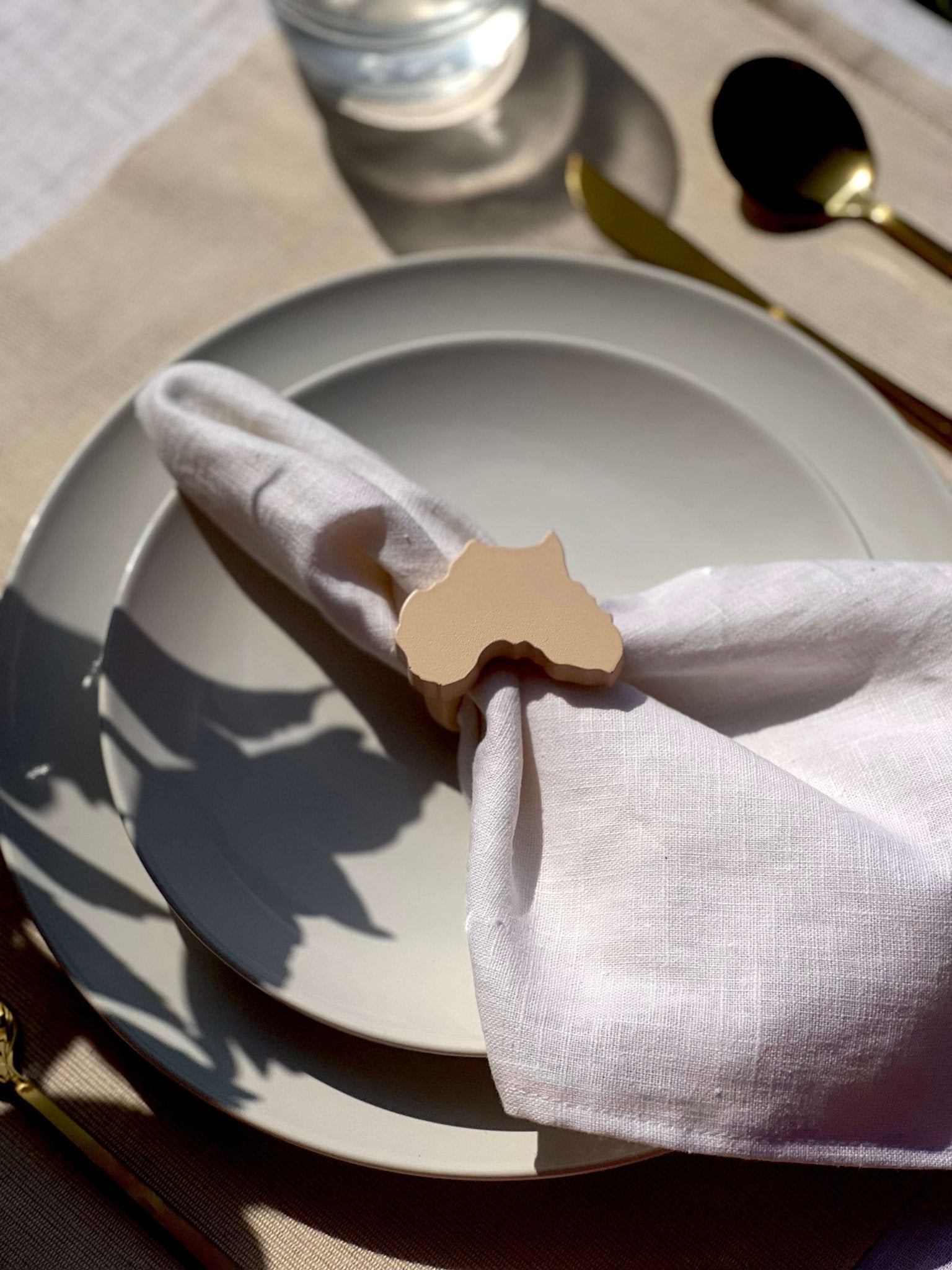 Napkin folded in a cream Africa-shaped napkin ring on an off white plate, gold cutlery with glass in the background.