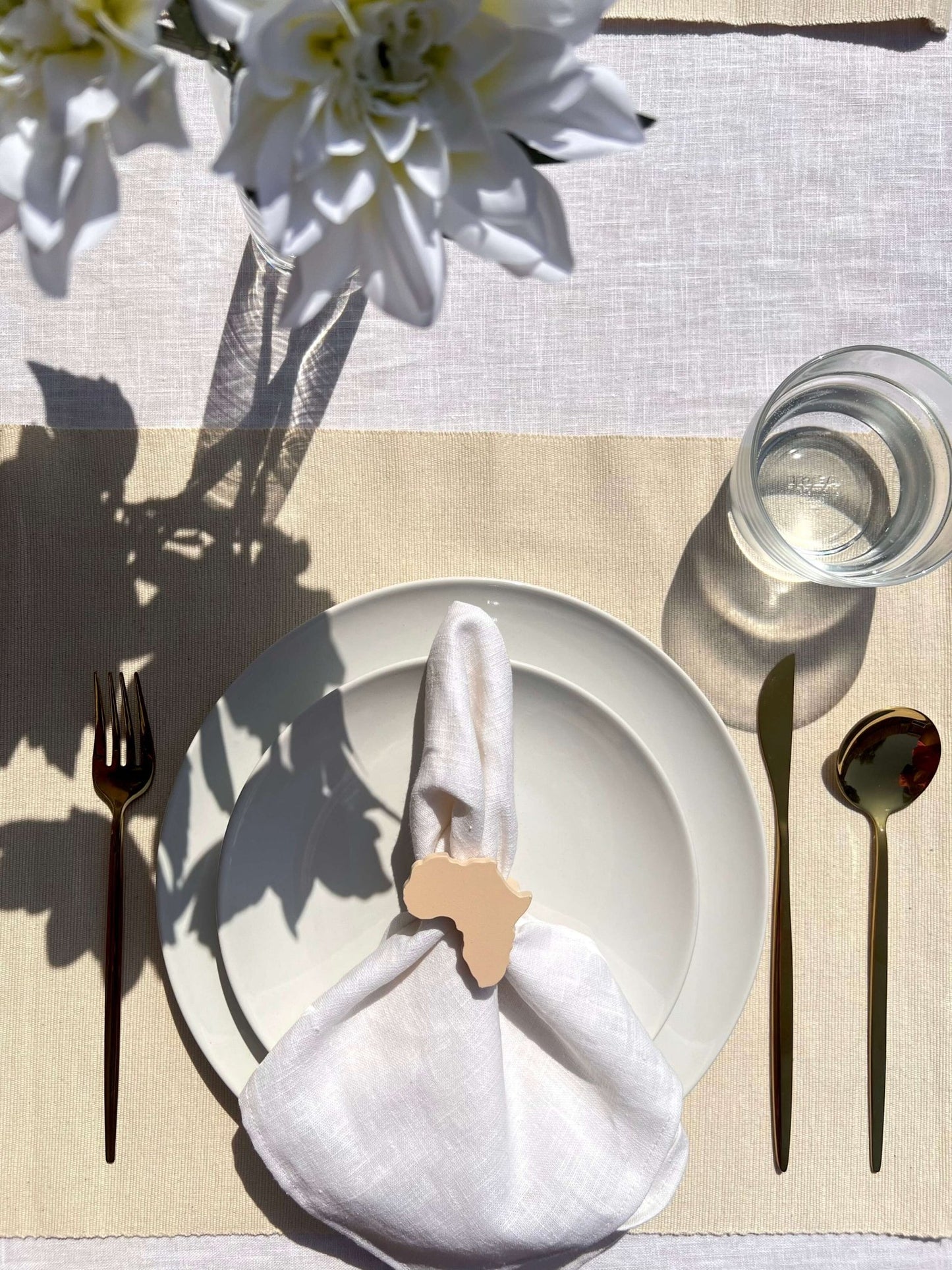 Dining table setting with off white plate, folded napkin with an Africa shaped napkin ring, gold cutlery and a glass of water on a beige tablecloth.