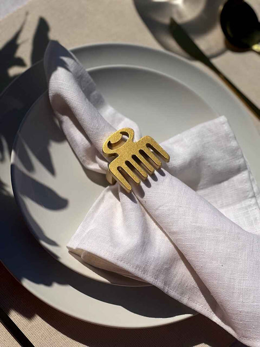 Dining table setting with off white plate, folded napkin with a gold Duafe adinkra napkin ring, gold cutlery and a glass of water on a beige tablecloth.