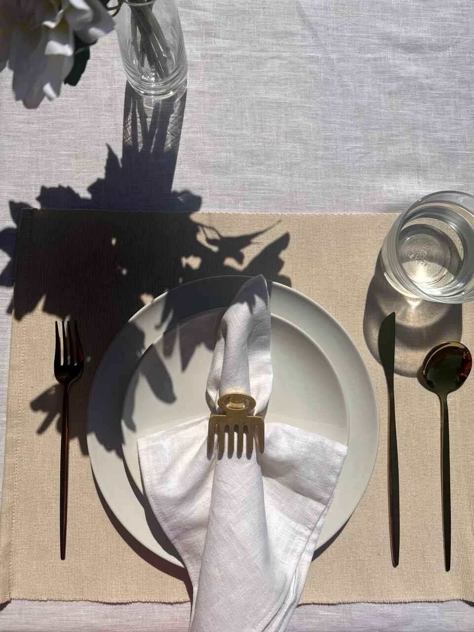 Dining table setting with off white plate, folded napkin with a gold Duafe adinkra napkin ring, gold cutlery and a glass of water on a beige tablecloth.