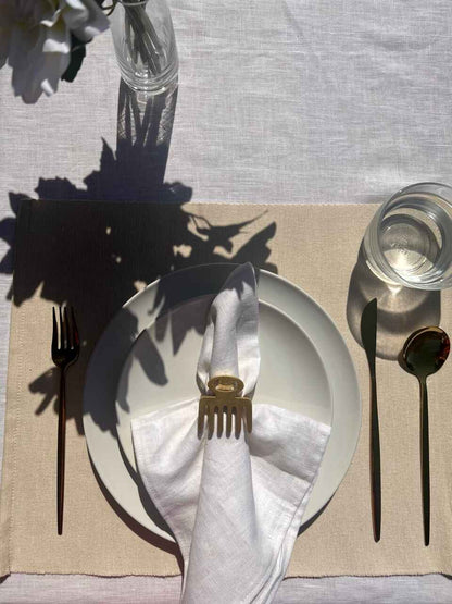 Dining table setting with off white plate, folded napkin with a gold Duafe adinkra napkin ring, gold cutlery and a glass of water on a beige tablecloth.