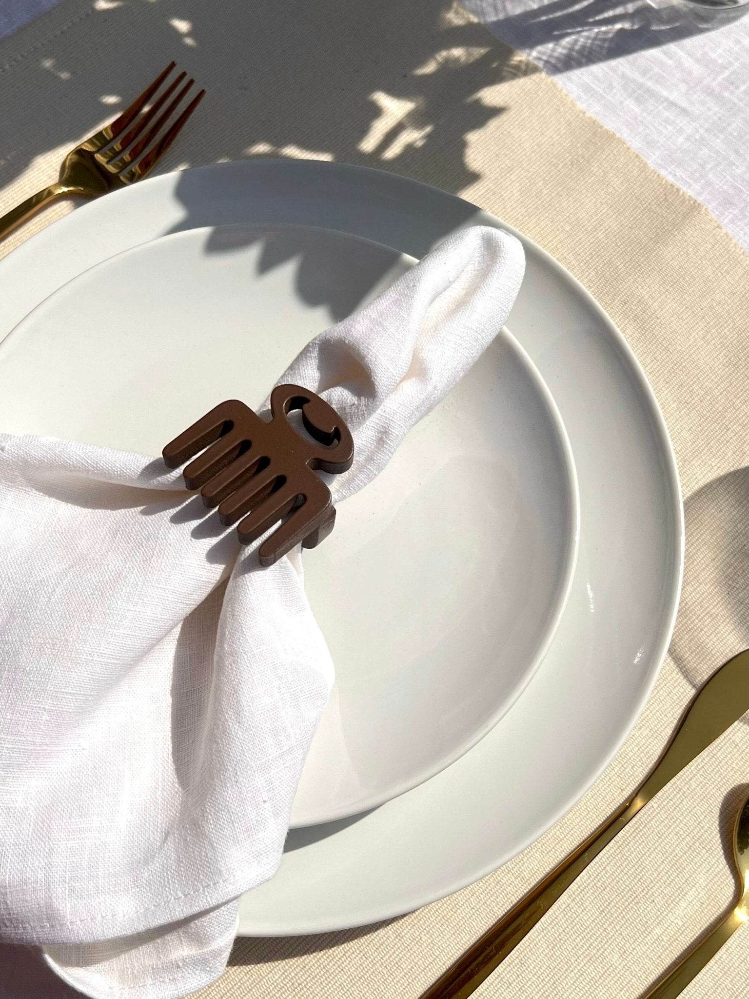 White napkin with a coffee coloured Duafe adinkra napkin ring on an off white plate, gold cutlery, and clear glass of water.