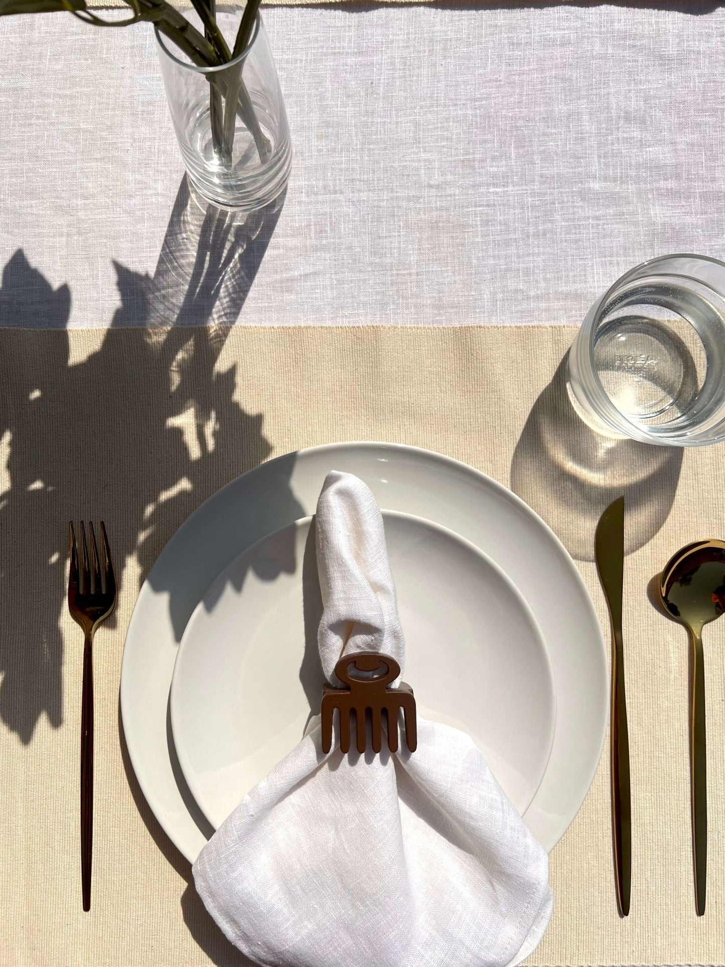 White napkin with a coffee coloured Duafe adinkra napkin ring on an off white plate, gold cutlery, and clear glass of water.