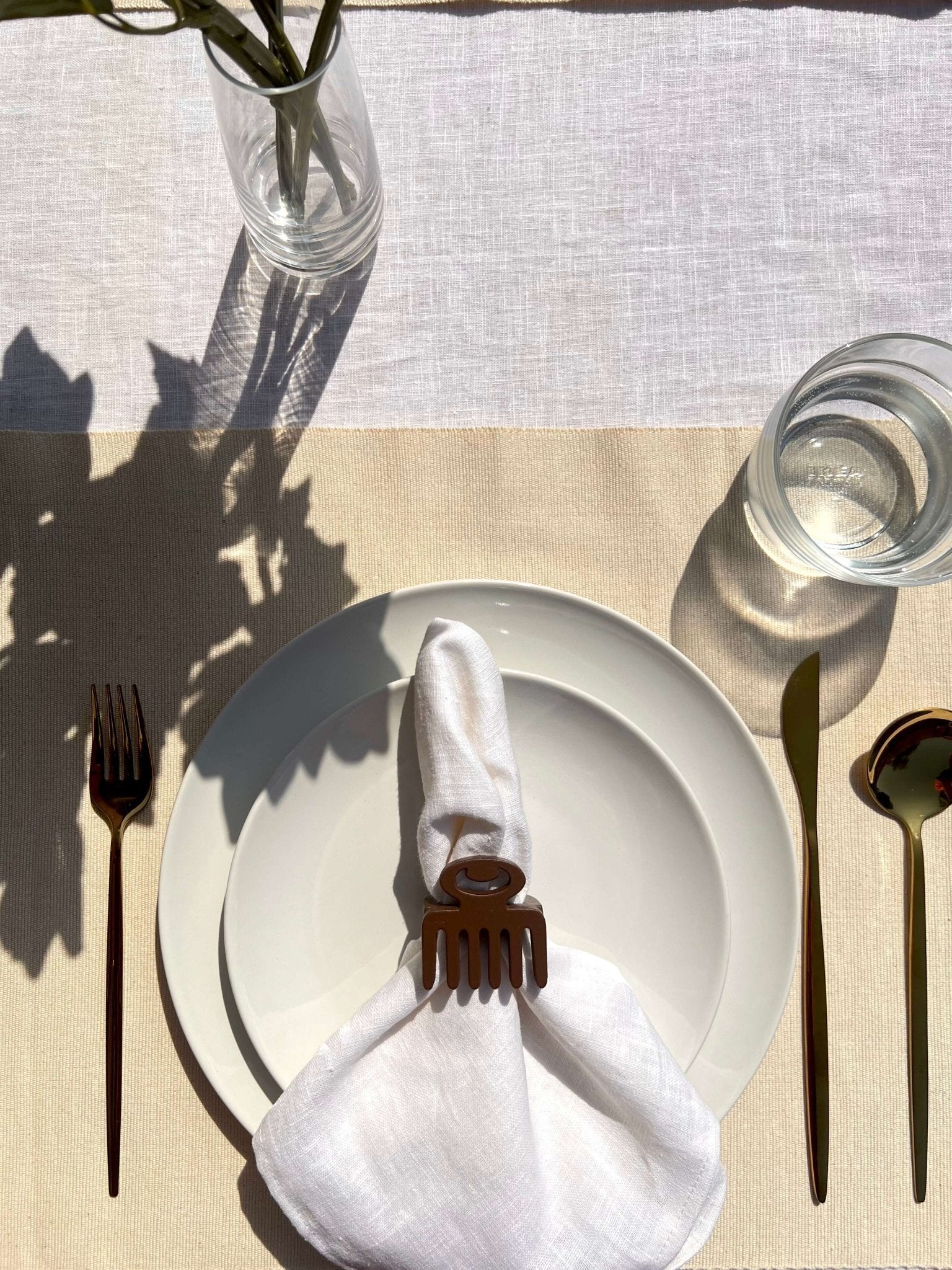 White napkin with a coffee coloured Duafe adinkra napkin ring on an off white plate, gold cutlery, and clear glass of water.