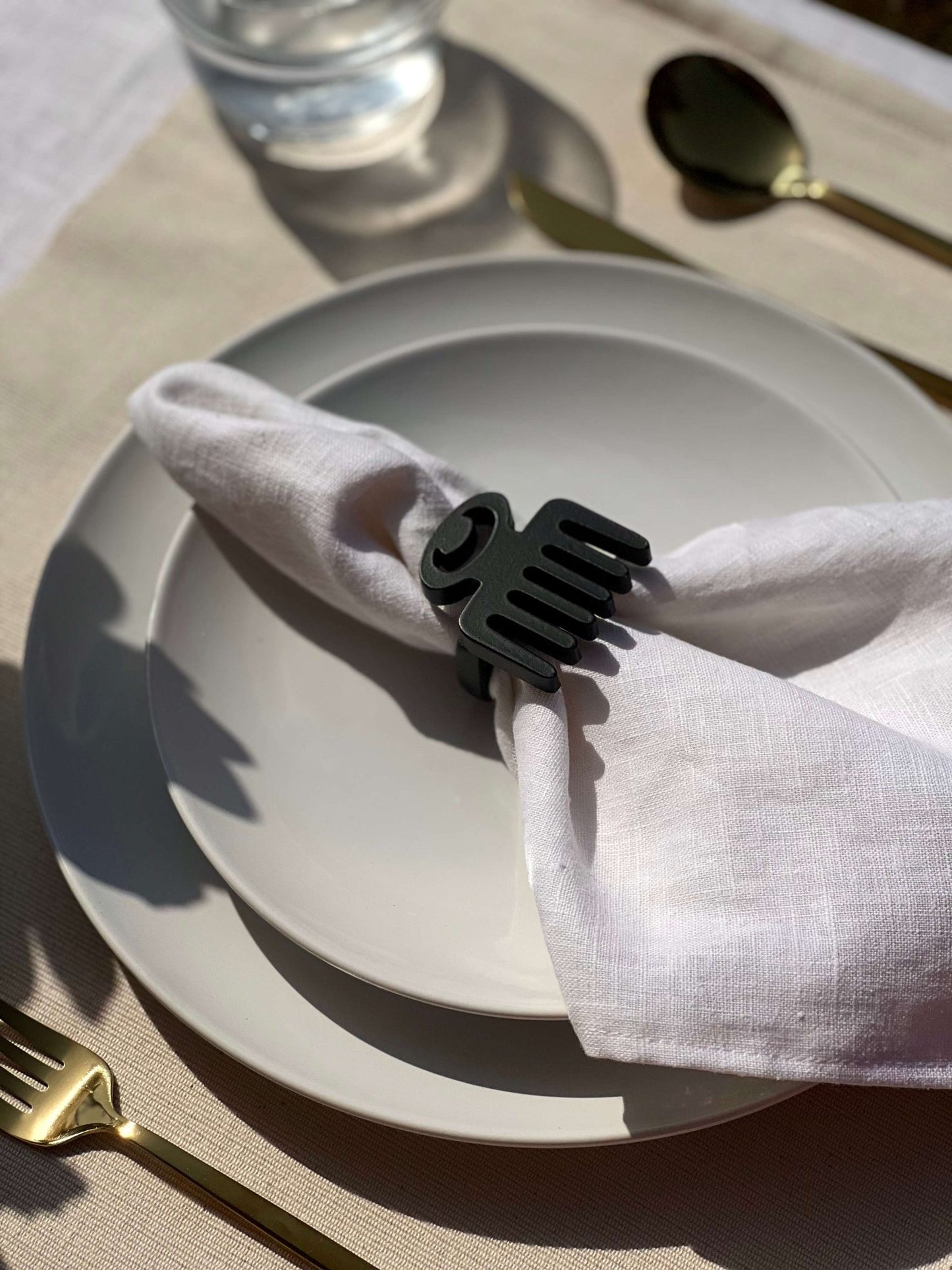  White napkin with a black Duafe adinkra napkin ring on an off white plate, gold cutlery, and clear glass of water.