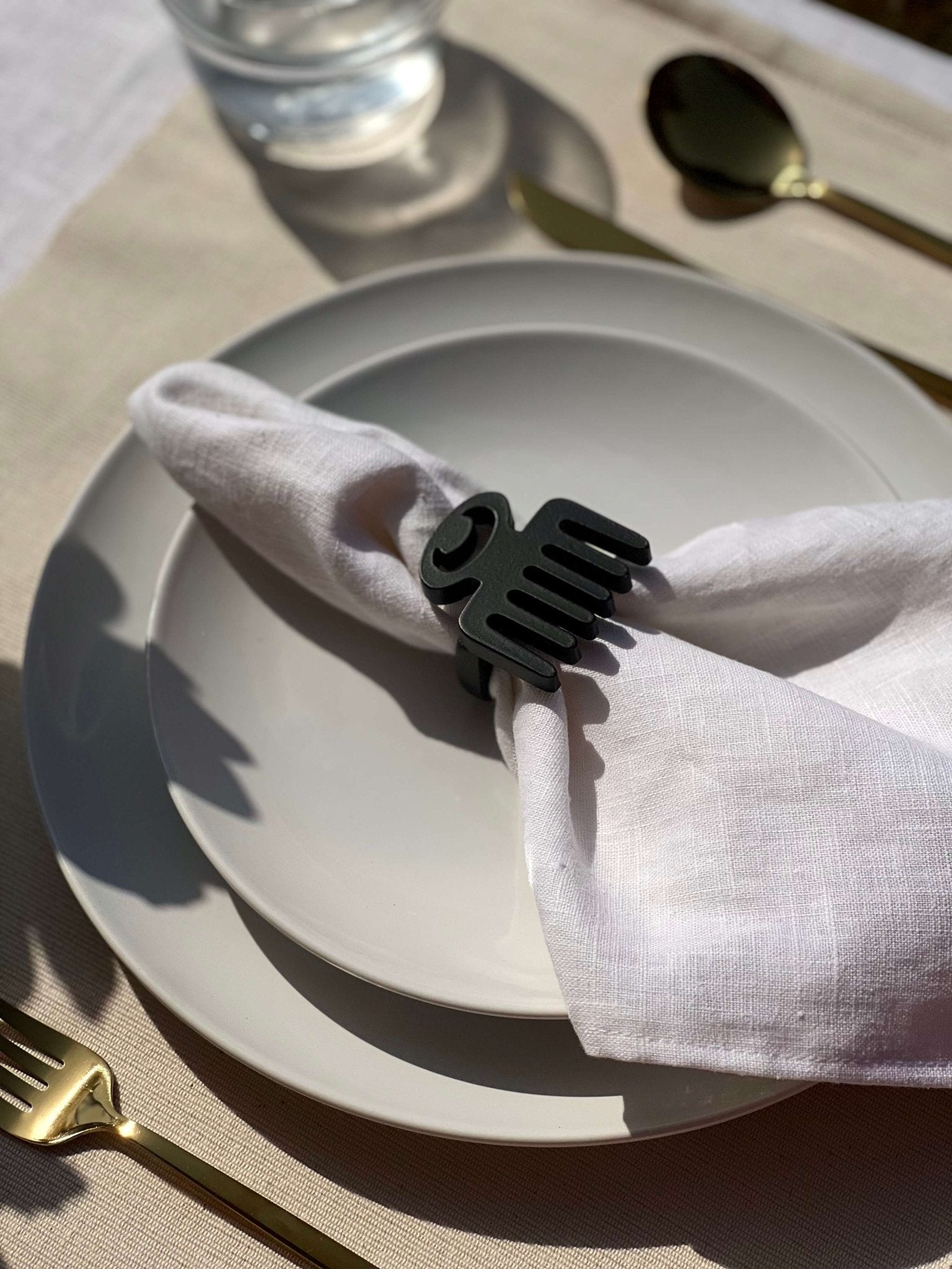  White napkin with a black Duafe adinkra napkin ring on an off white plate, gold cutlery, and clear glass of water.