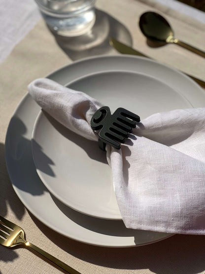  White napkin with a black Duafe adinkra napkin ring on an off white plate, gold cutlery, and clear glass of water.