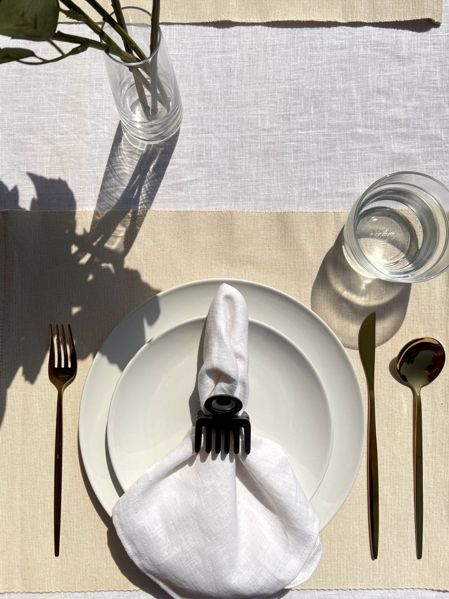  White napkin with a black Duafe adinkra napkin ring on an off white plate, gold cutlery, and clear glass of water.