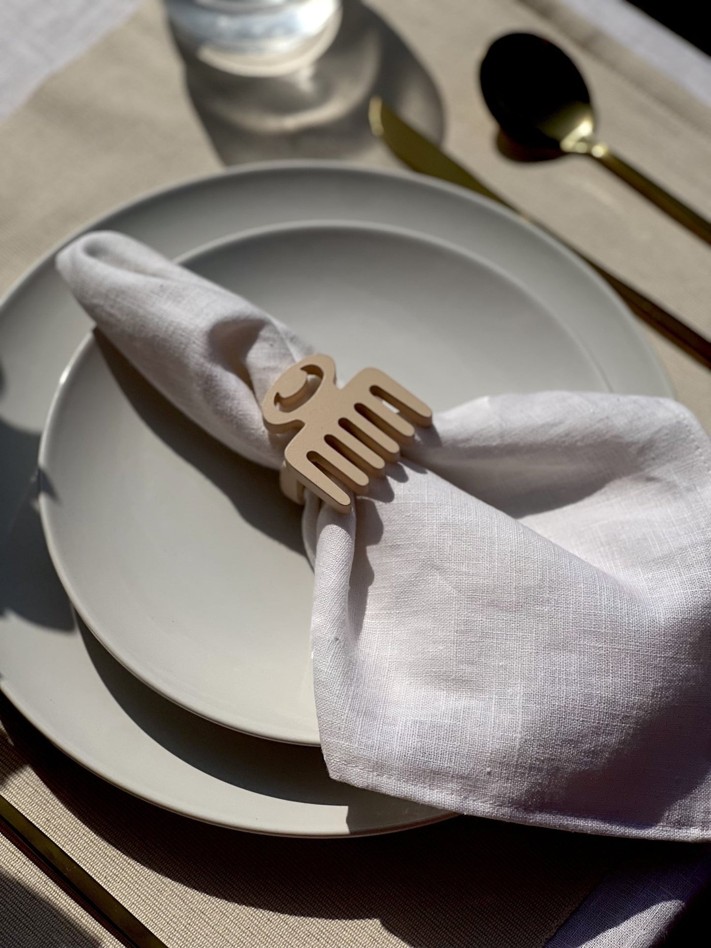 White napkin with a cream Duafe adinkra napkin ring on an off white plate, gold cutlery, and a clear glass of water.
