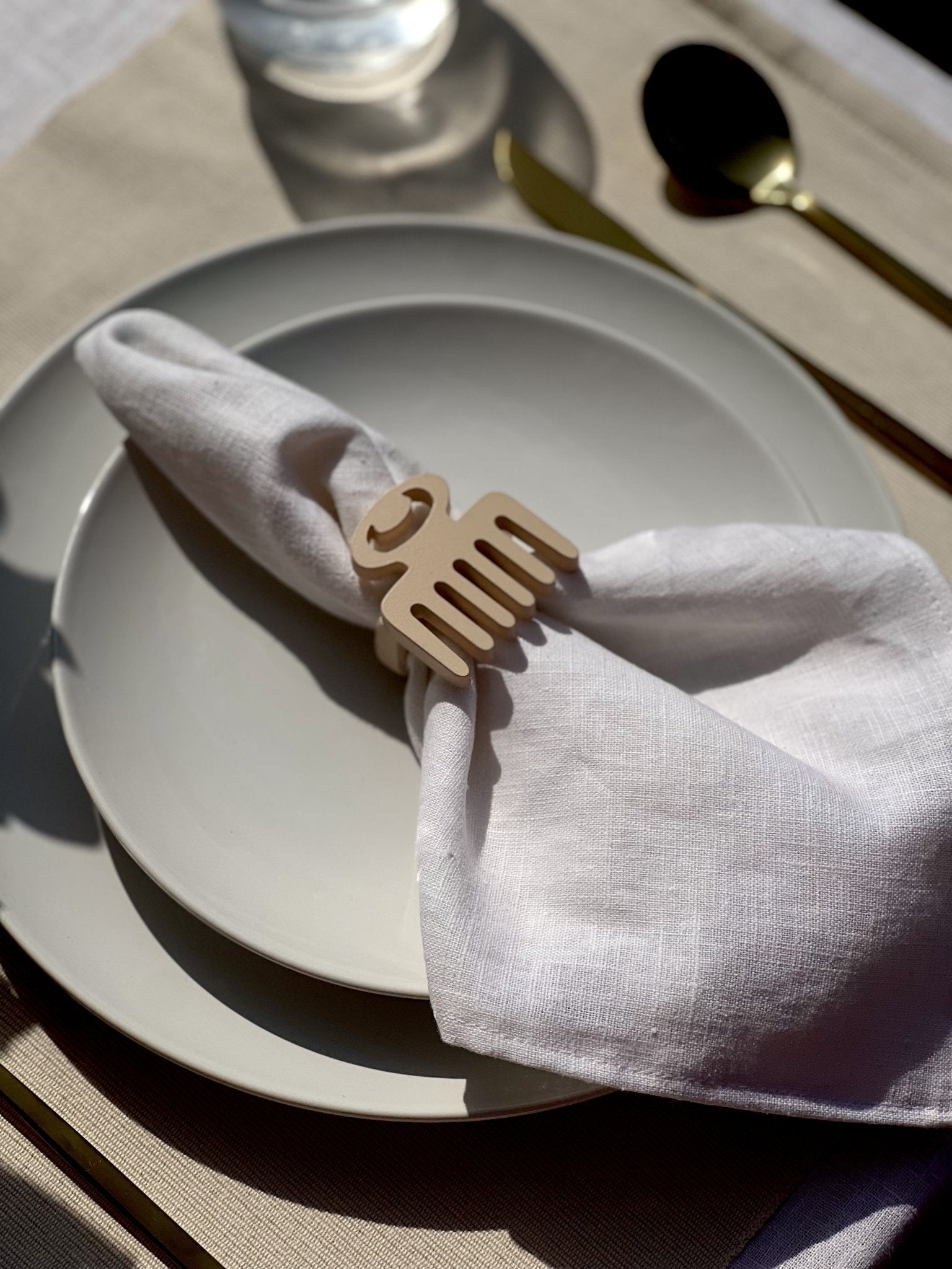 White napkin with a cream Duafe adinkra napkin ring on an off white plate, gold cutlery, and a clear glass of water.