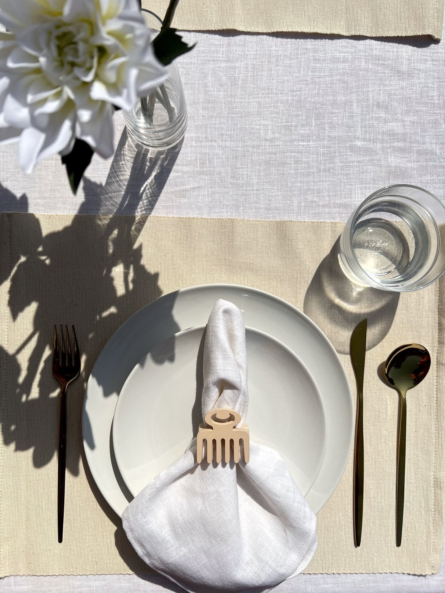 White napkin with a cream Duafe adinkra napkin ring on an off white plate, gold cutlery, and a clear glass of water.