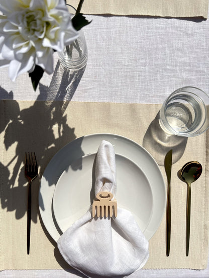 White napkin with a cream Duafe adinkra napkin ring on an off white plate, gold cutlery, and a clear glass of water.