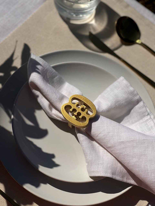 A gold coloured Ese Ne Tekrema adinkra napkin ring on an off white plate, glass of water and gold cutlery.
