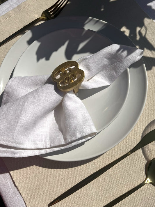 A gold coloured Ese Ne Tekrema adinkra napkin ring on an off white plate, glass of water and gold cutlery.