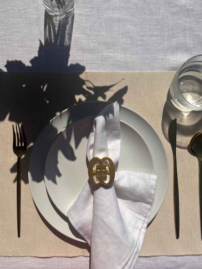 A gold coloured Ese Ne Tekrema adinkra napkin ring on an off white plate, glass of water and gold cutlery.