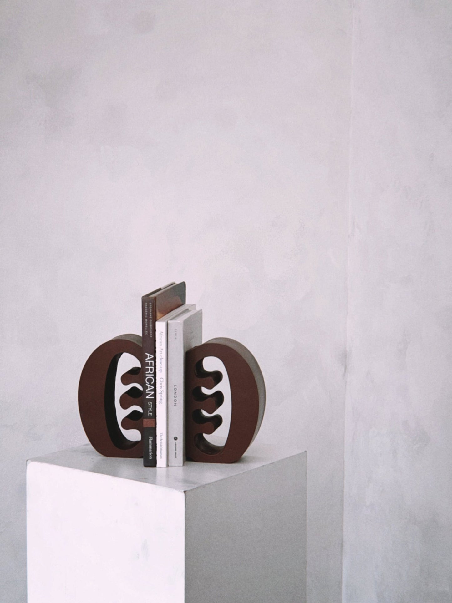 Decorative coffee coloured Ese Ne Tekrema adinkra bookends on a white plinth with a light gray lime wash background.