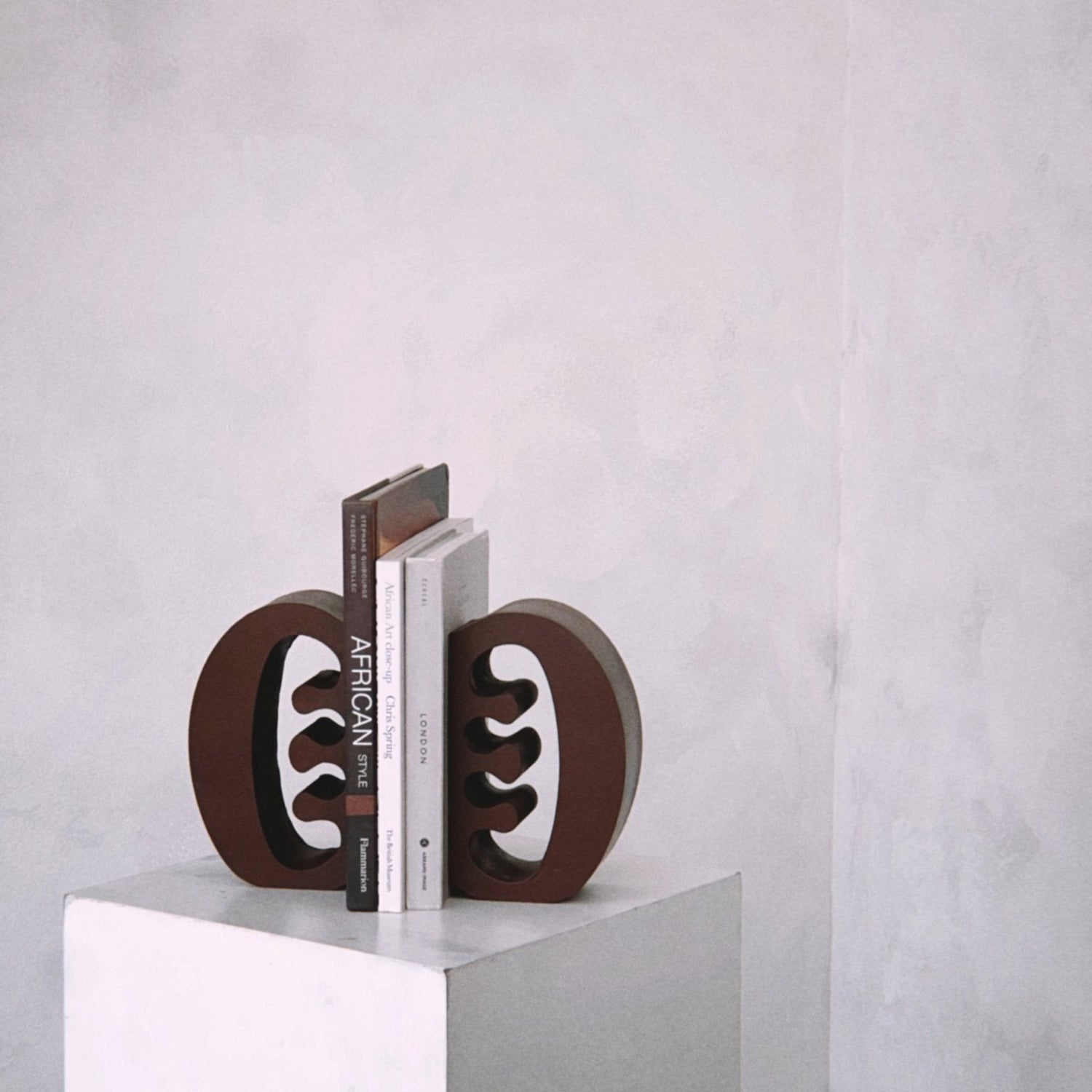Decorative coffee coloured Ese Ne Tekrema adinkra bookends on a white plinth with a light gray lime wash background.