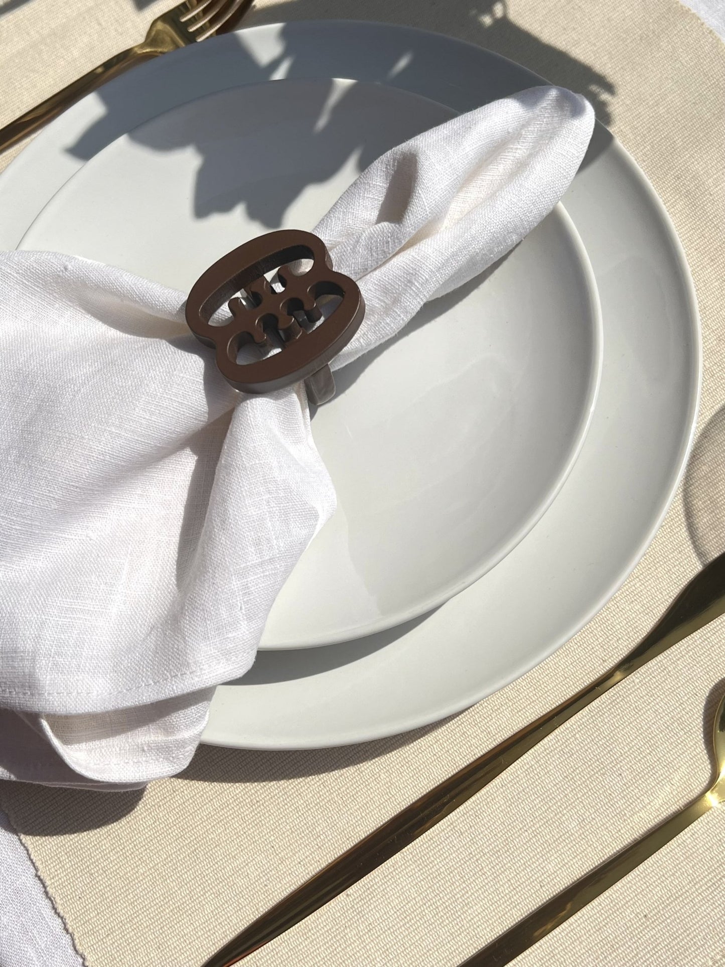  A coffee coloured Ese Ne Tekrema adinkra napkin ring on an off white plate, with gold cutlery.