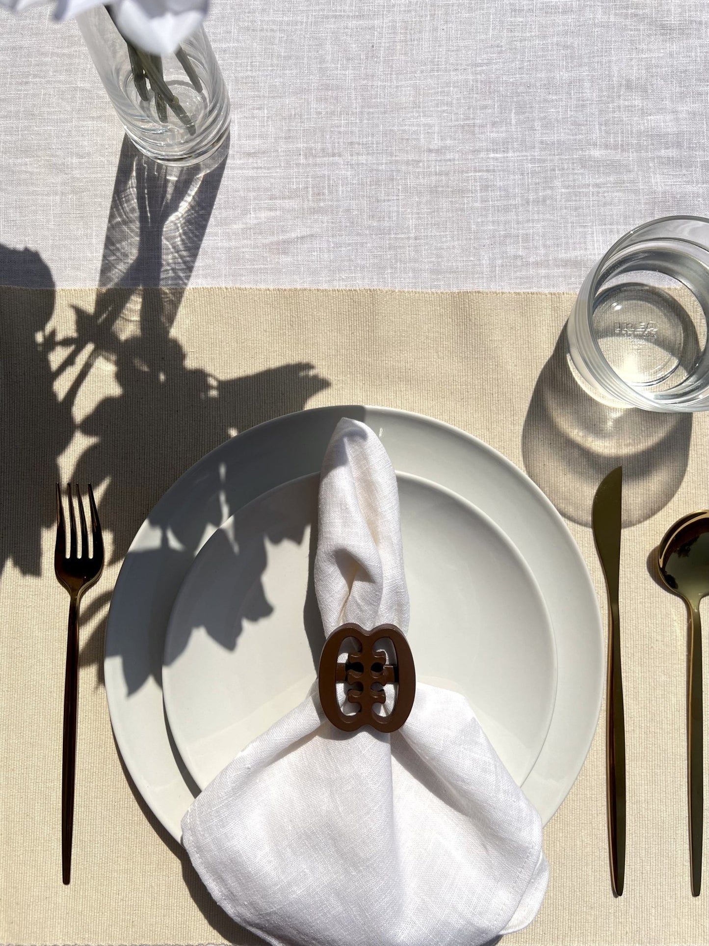  A coffee coloured Ese Ne Tekrema adinkra napkin ring on an off white plate, with gold cutlery.