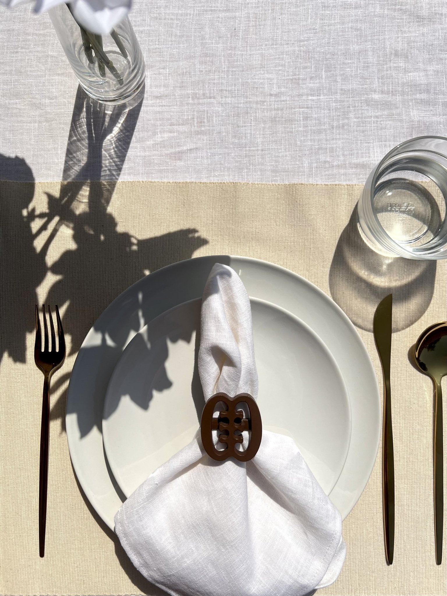  A coffee coloured Ese Ne Tekrema adinkra napkin ring on an off white plate, with gold cutlery.