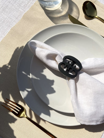 Ese Ne Tekrema napkin ring in black on an off white plate, glass of water and gold cutlery, on a cream placemat.