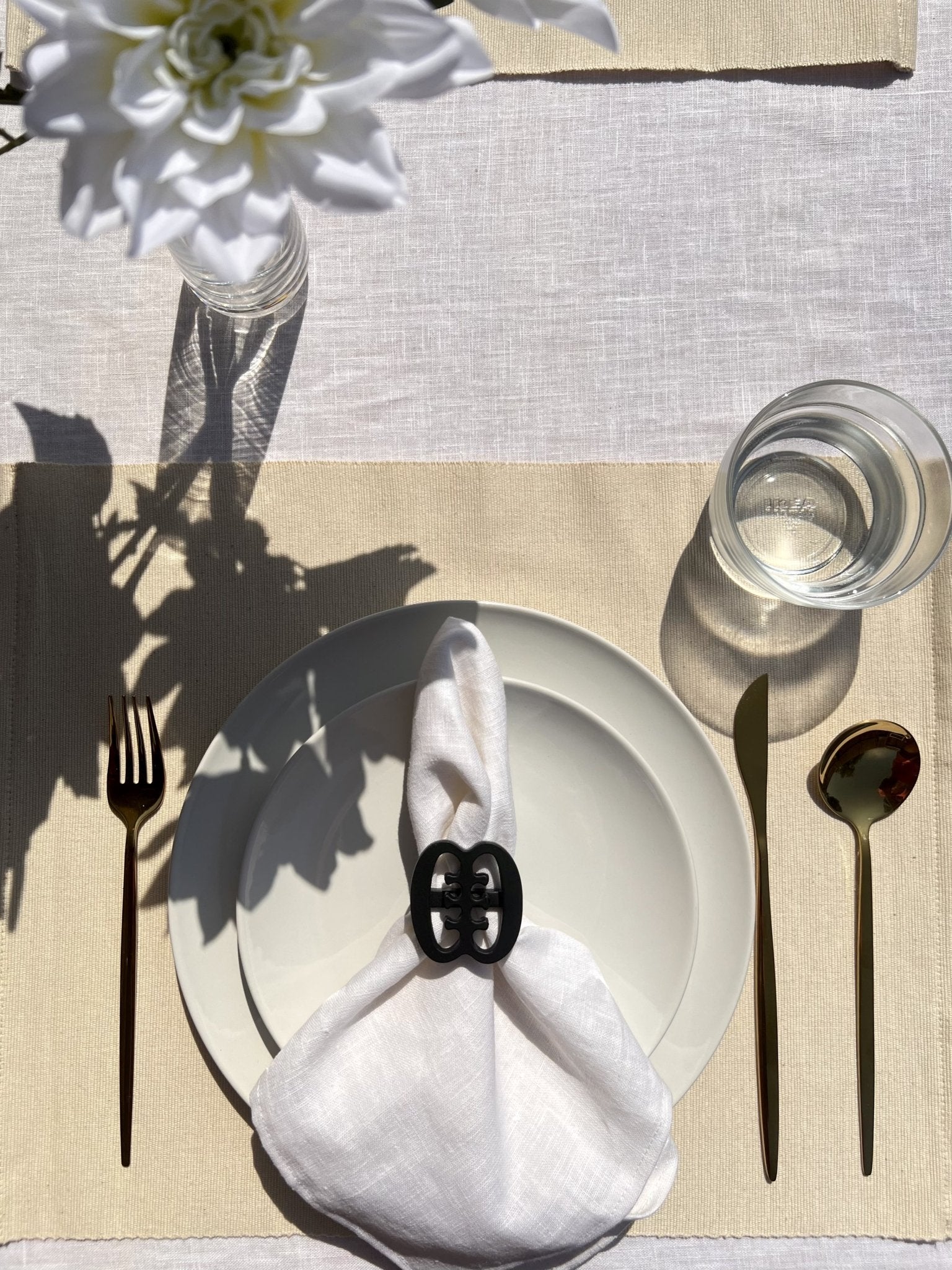 Ese Ne Tekrema napkin ring in black on an off white plate, glass of water and gold cutlery, on a cream placemat.