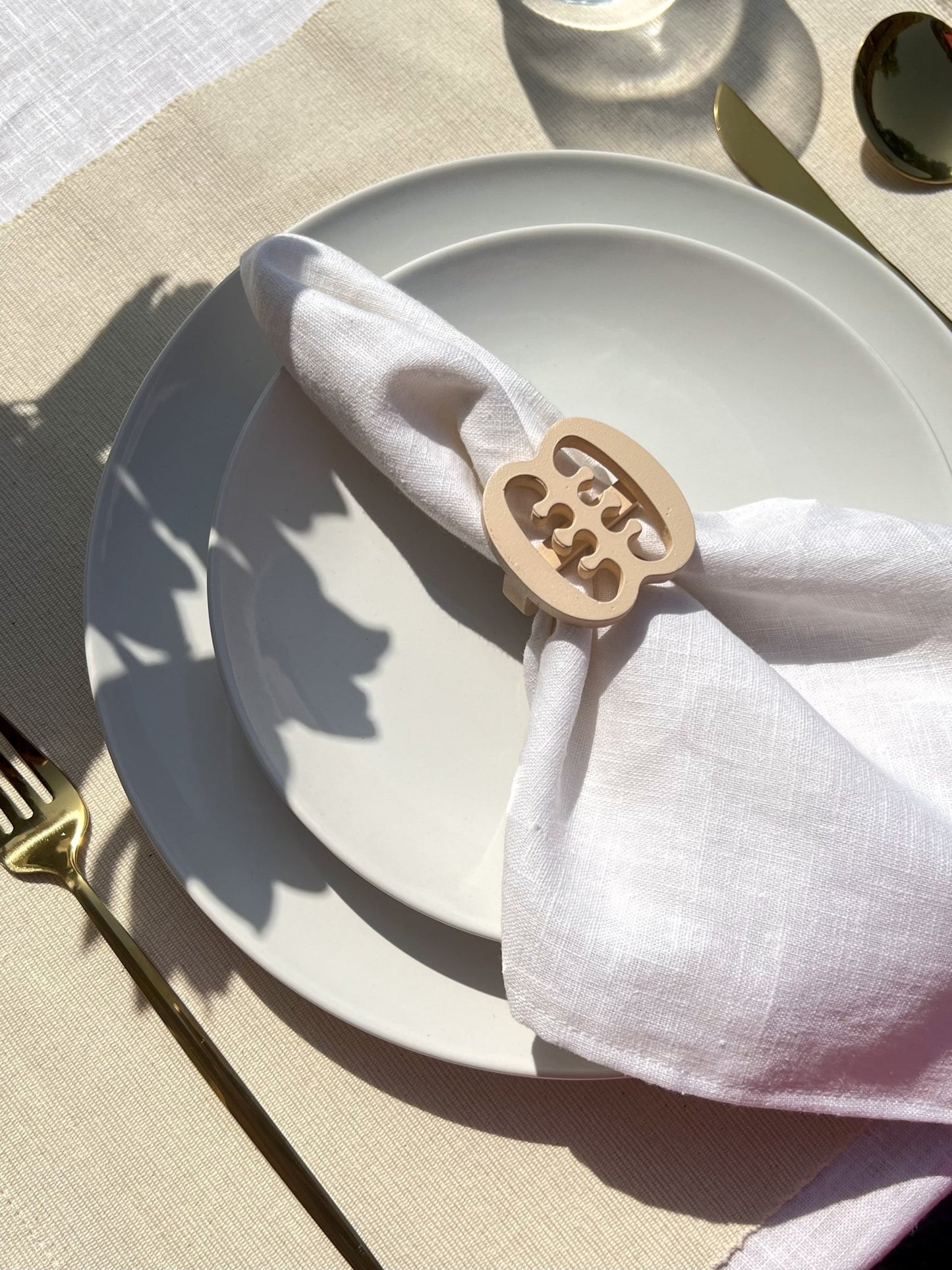 White napkin with a cream Ese Ne Tekrema adinkra napkin ring on an off white plate, gold cutlery, and a clear glass of water.