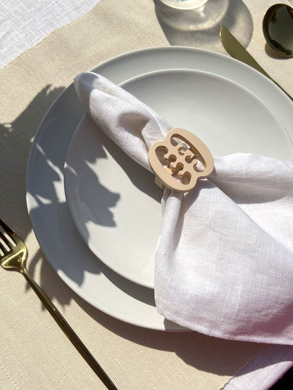 White napkin with a cream Ese Ne Tekrema adinkra napkin ring on an off white plate, gold cutlery, and a clear glass of water.