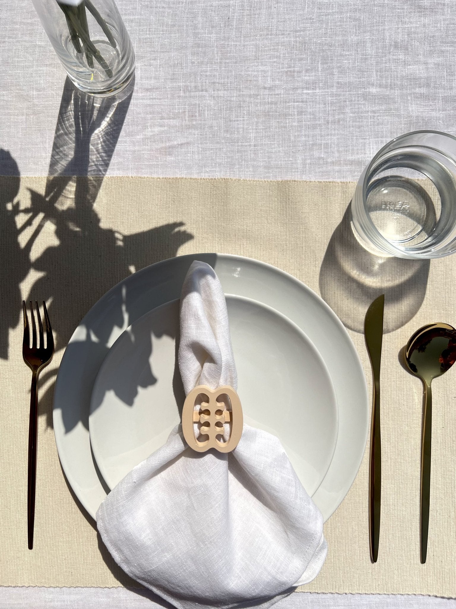 White napkin with a cream Ese Ne Tekrema adinkra napkin ring on an off white plate, gold cutlery, and a clear glass of water.