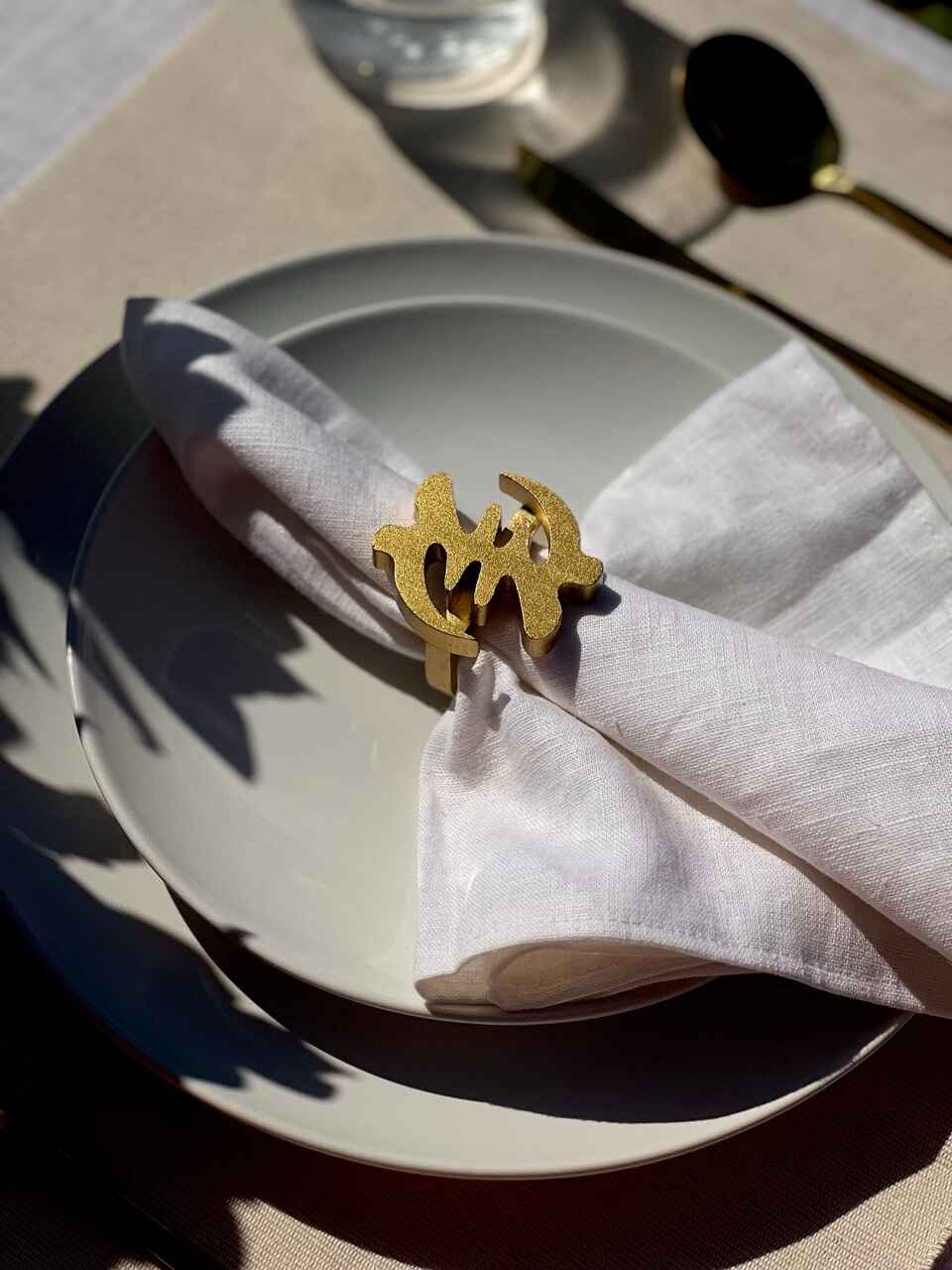 White napkin with a gold Gye Nyame adinkra napkin ring on an off white plate, gold cutlery, and clear glass of water.