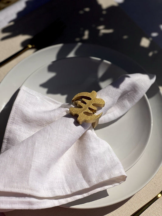 White napkin with a gold Gye Nyame adinkra napkin ring on an off white plate, gold cutlery, and clear glass of water.