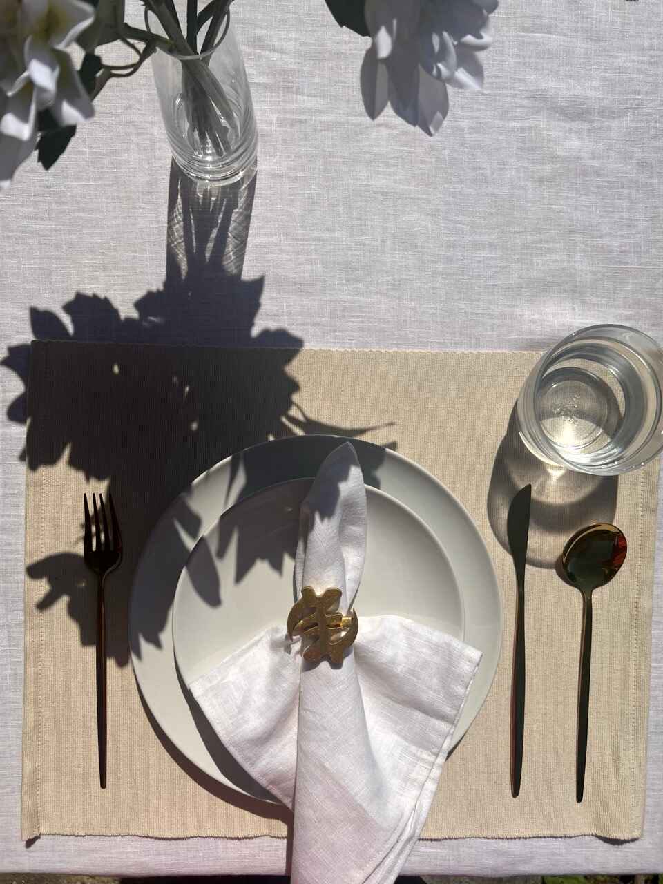 White napkin with a gold Gye Nyame adinkra napkin ring on an off white plate, gold cutlery, and clear glass of water.