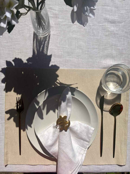 White napkin with a gold Gye Nyame adinkra napkin ring on an off white plate, gold cutlery, and clear glass of water.