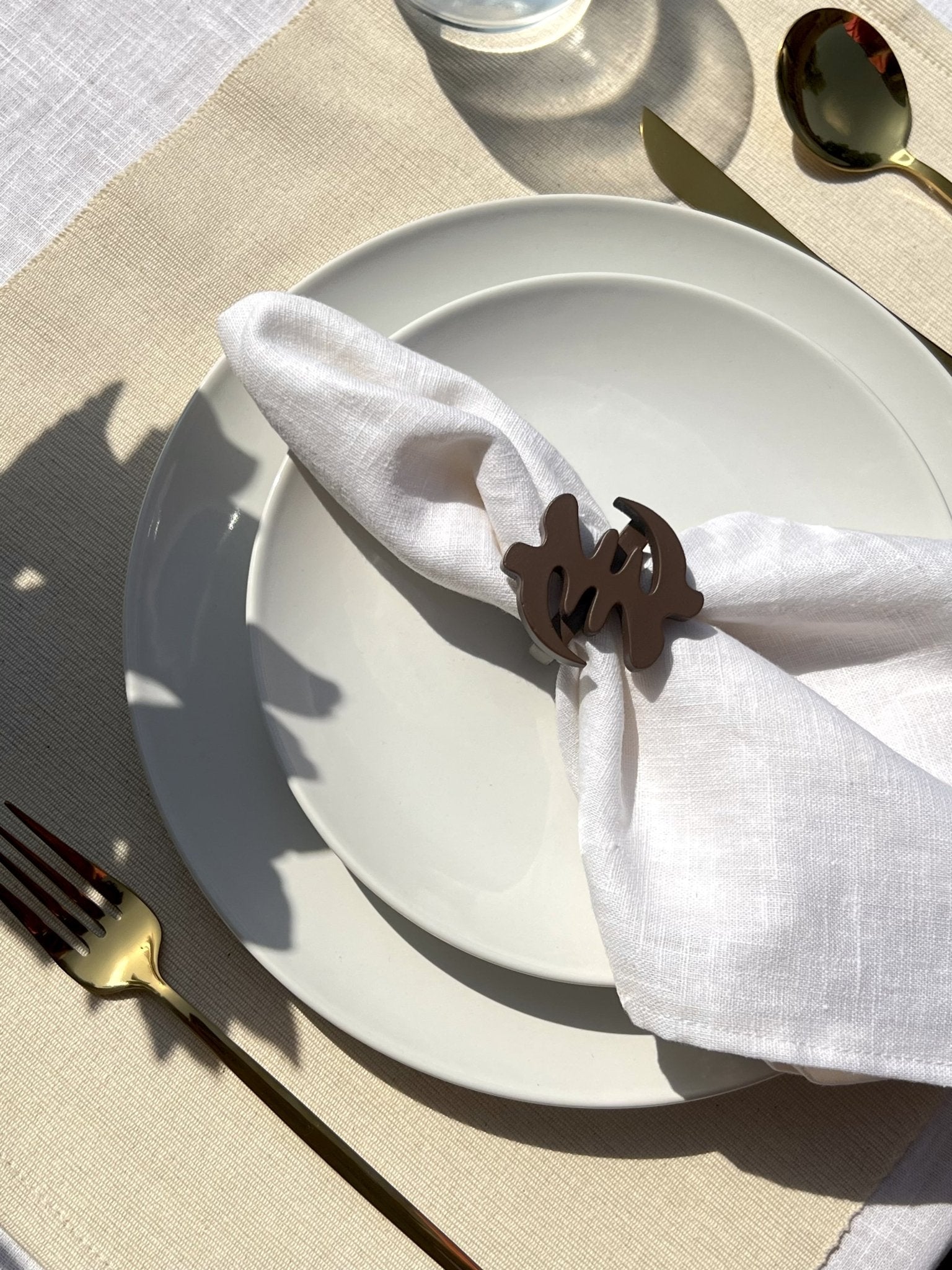 White napkin with a coffee coloured Gye Nyame adinkra napkin ring on an off white plate with gold cutlery.