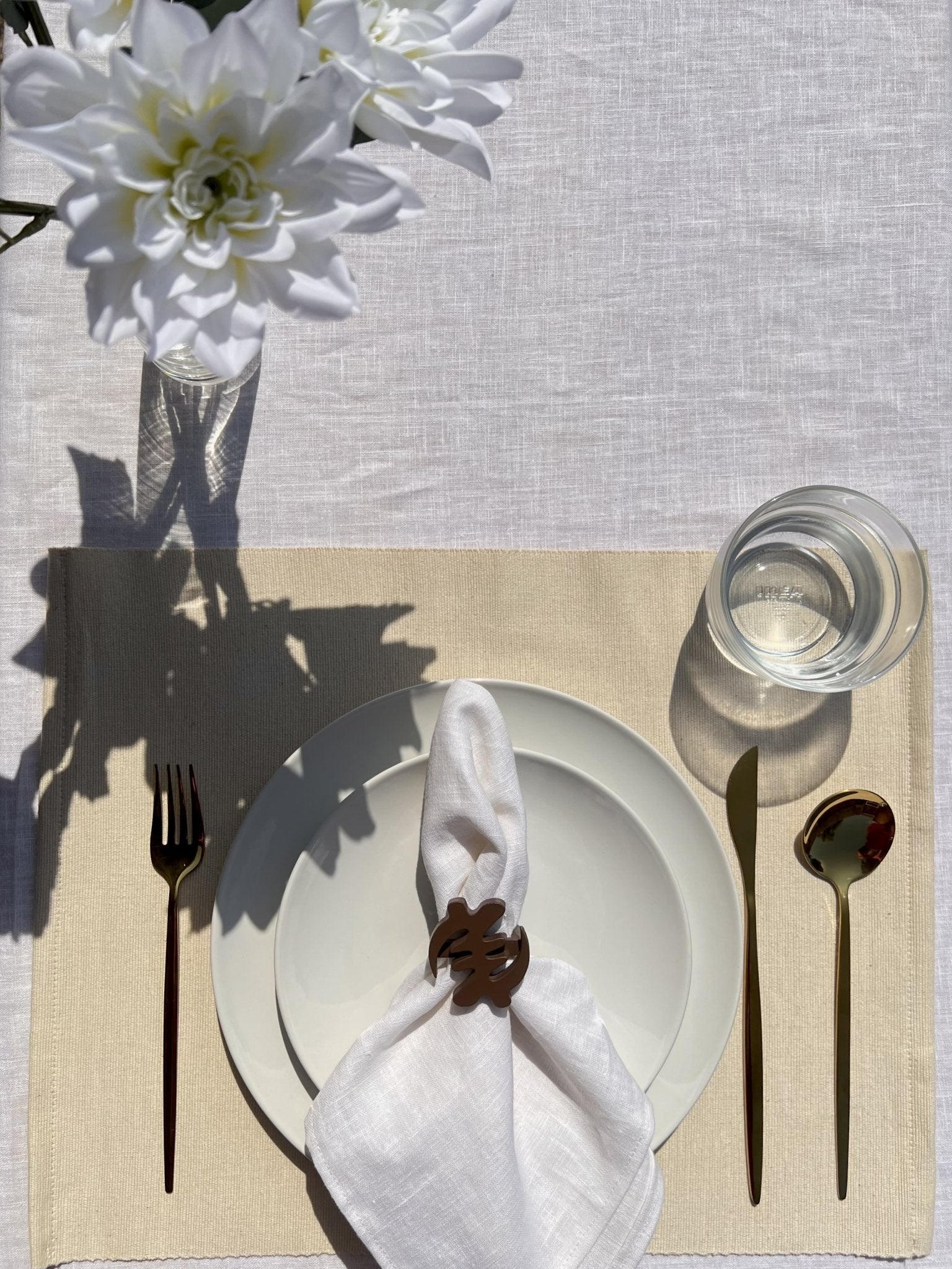 White napkin with a coffee coloured Gye Nyame adinkra napkin ring on an off white plate with gold cutlery.