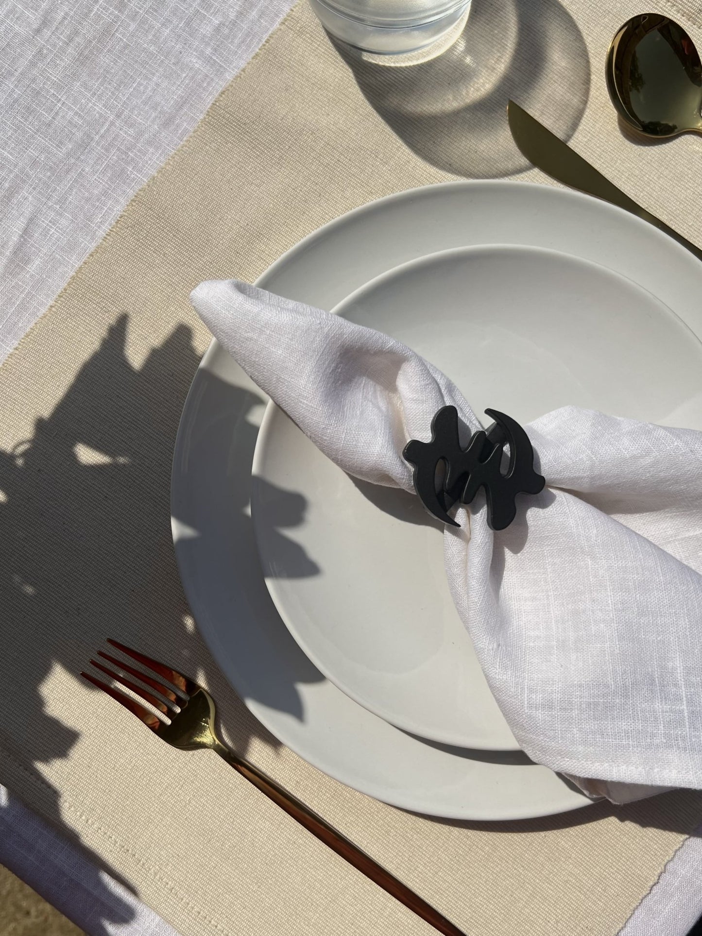 White napkin with a black Gye Nyame adinkra napkin ring on an off white plate, gold cutlery, and clear glass of water.

