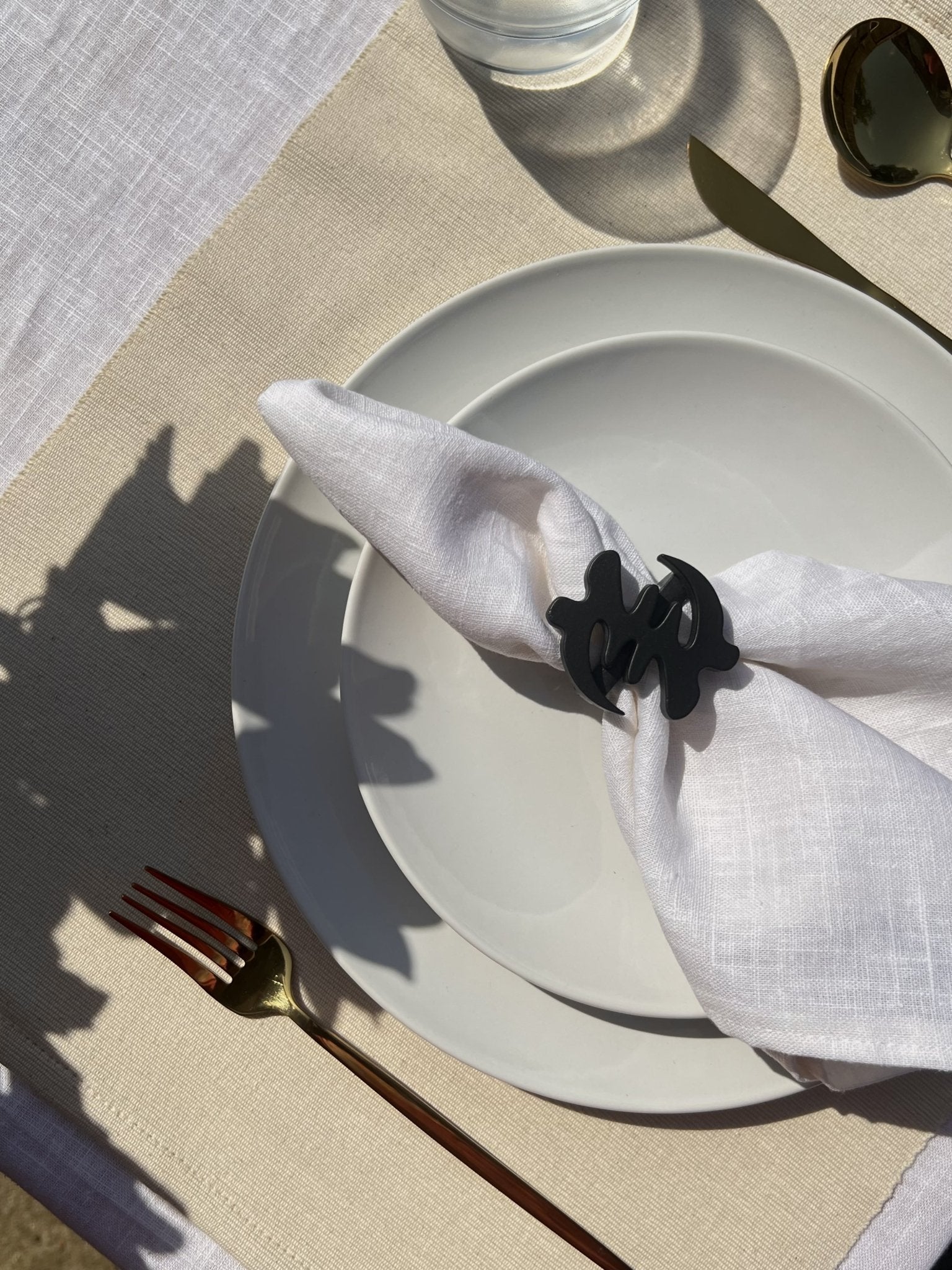 White napkin with a black Gye Nyame adinkra napkin ring on an off white plate, gold cutlery, and clear glass of water.
