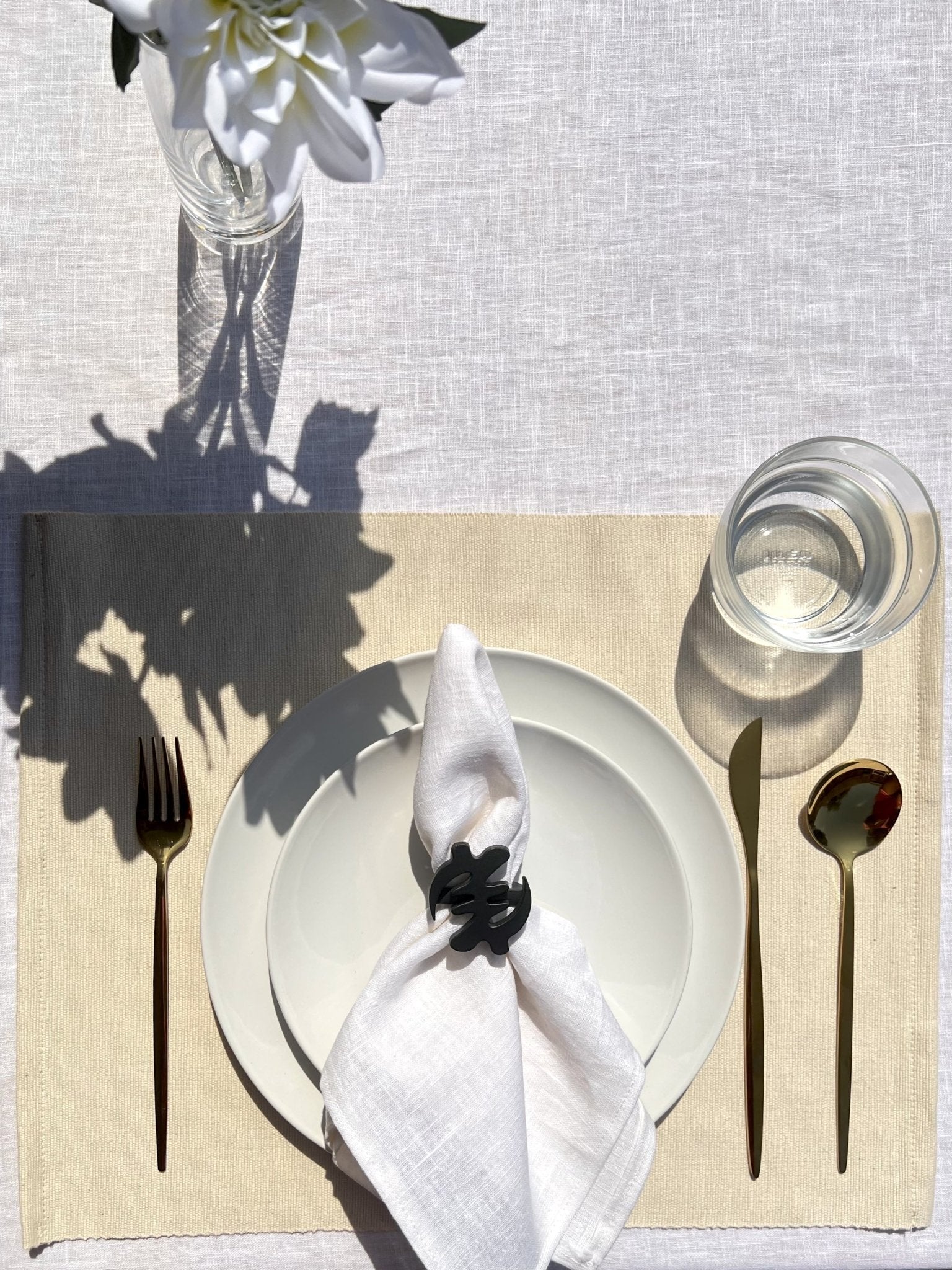 White napkin with a black Gye Nyame adinkra napkin ring on an off white plate, gold cutlery, and clear glass of water.
