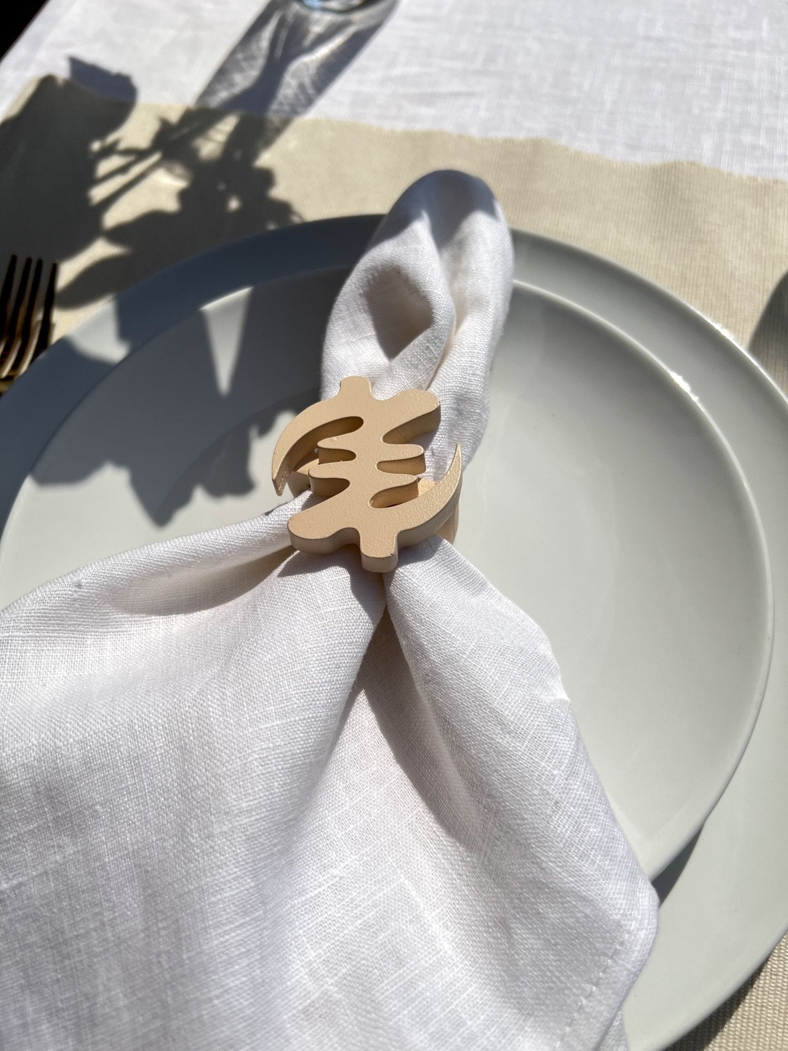 Napkin folded in a cream Gye Nyame adinkra napkin ring on an off white plate with gold cutlery 